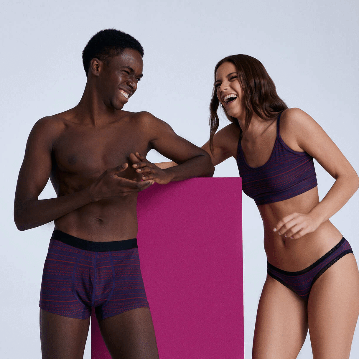 Two smiling models in matching plum organic cotton underwear from Albero Natur — a man in boxer briefs and a woman in a bralette and briefs — leaning on a magenta block against a pale lavender background.