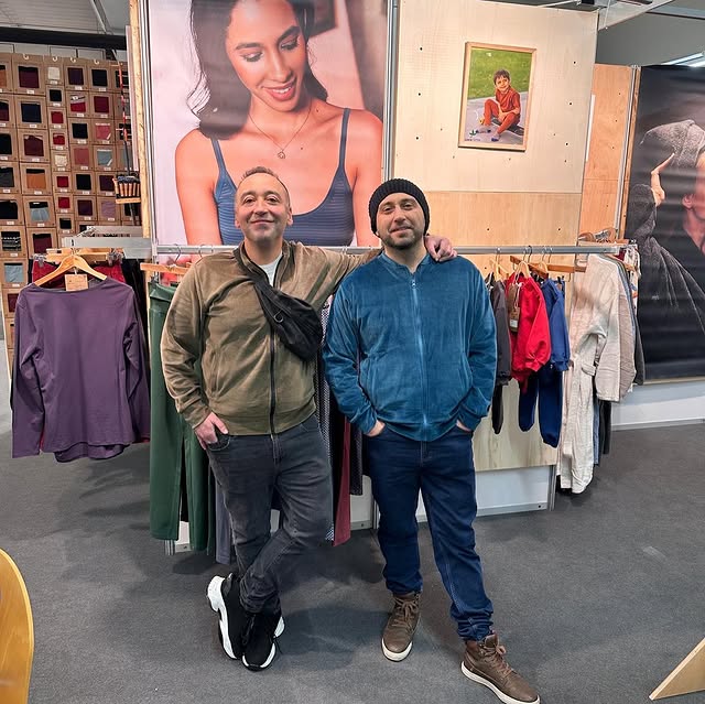 Two men posing together in a clothing store, with one wearing a green zip-up jacket and gray jeans, and the other wearing a blue zip-up jacket, dark jeans, and brown sneakers. Clothing racks and large wall posters are visible in the background.
