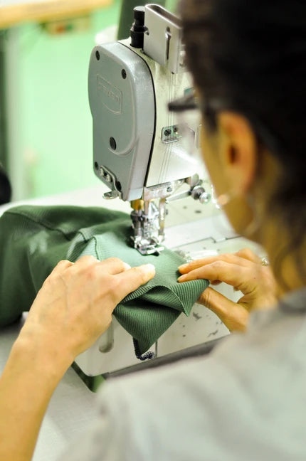 A close-up action shot of a person operating an industrial sewing machine, guiding a dark green piece of fabric under the needle. The person is seen from behind, wearing glasses and a light gray shirt, with the sewing machine head and metallic presser visible.